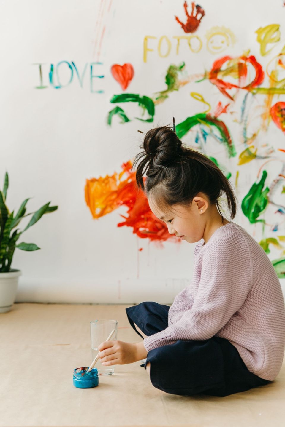 A girl sitting on the floor painting, with a wall covered in paintings behind her