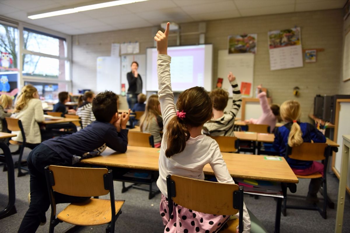 Children raising their hands in a classroom while the teacher stands at the front near a whiteboard