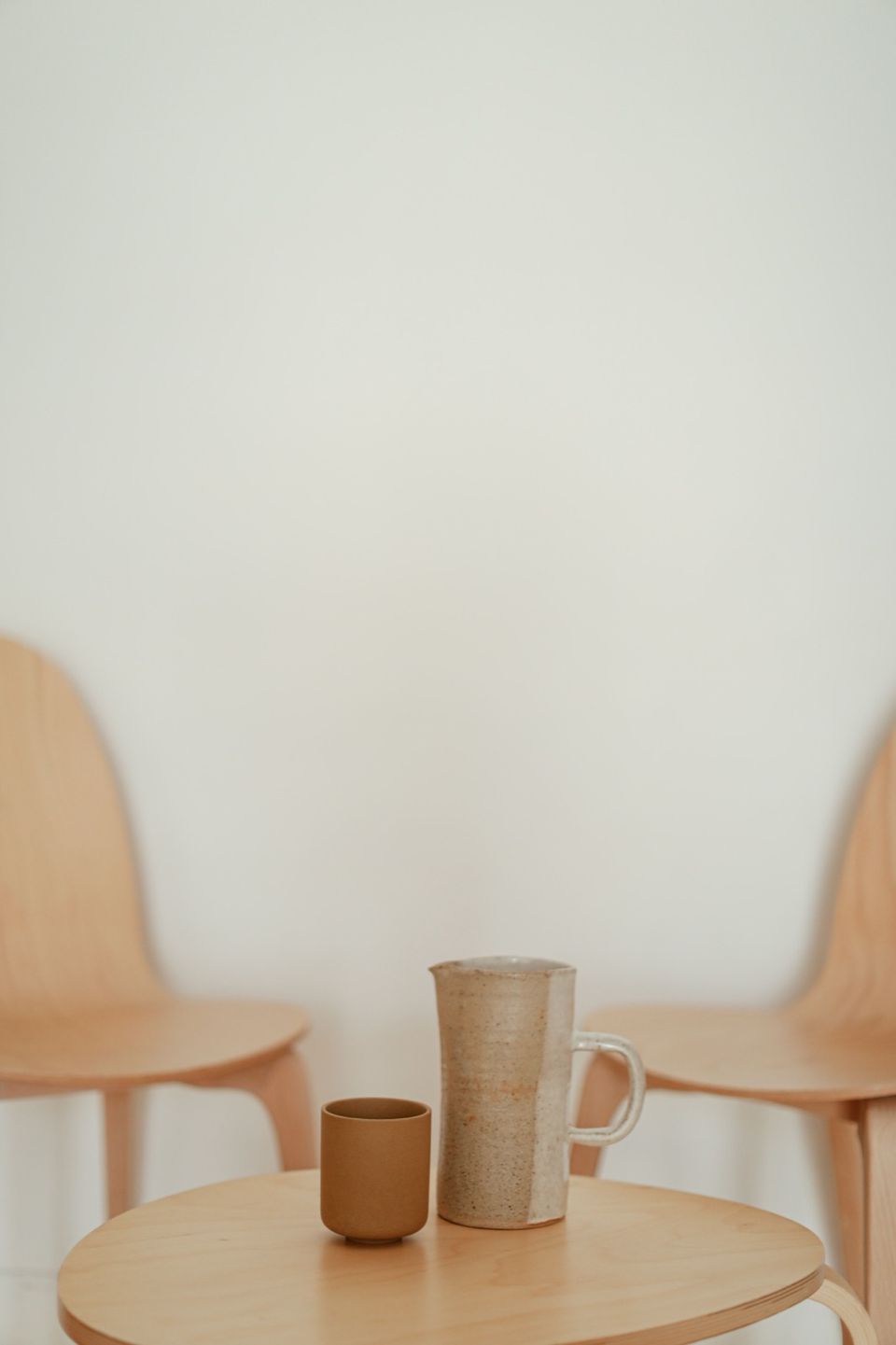 A ceramic pitcher and cup on a wooden table between two chairs in a bright, quiet room