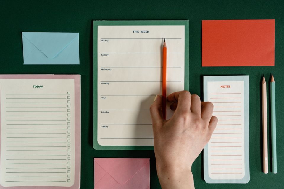 A hand writing on a weekly planner surrounded by notebooks and pencils